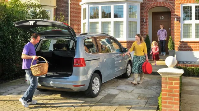 Blue Volkswagen Sharan parked out front of a house, with a family of four getting into the car. The boot is open, with one parent in a purple shirt placing a brown picnic basket in the boot.